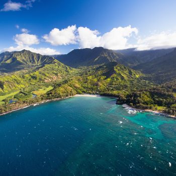 Coastal mountains rise above lush green landscape, surrounded by clear blue ocean under a sky with scattered clouds, creating a vibrant, natural scenery.