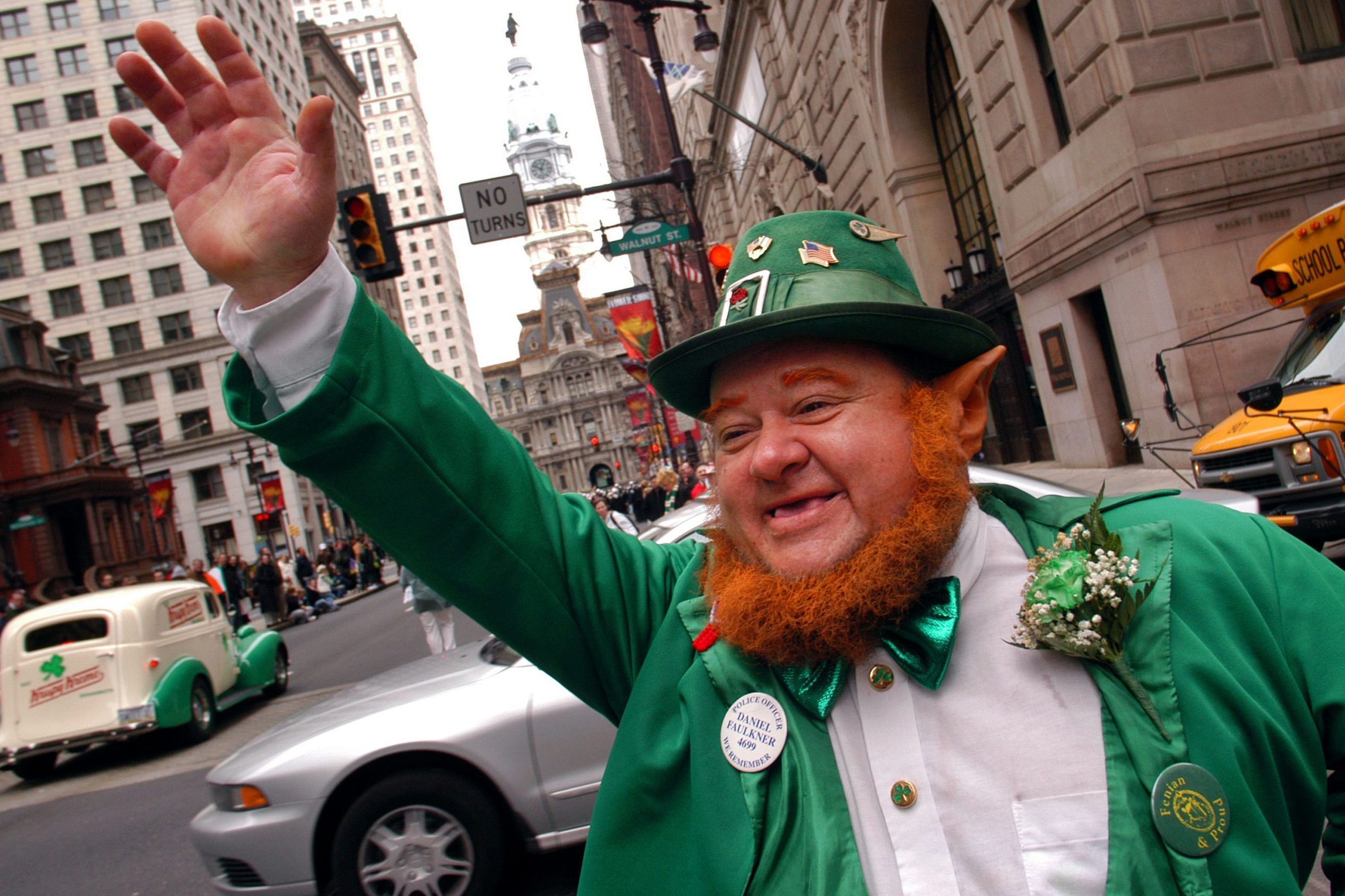 A man in a green outfit waves on a city street with tall buildings and vintage cars.