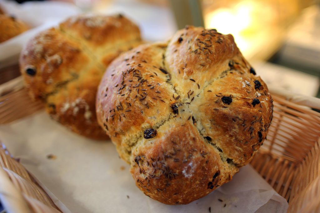 Round loaf of bread sits in a basket, sprinkled with seeds and raisins, surrounded by a warm, softly lit background.
