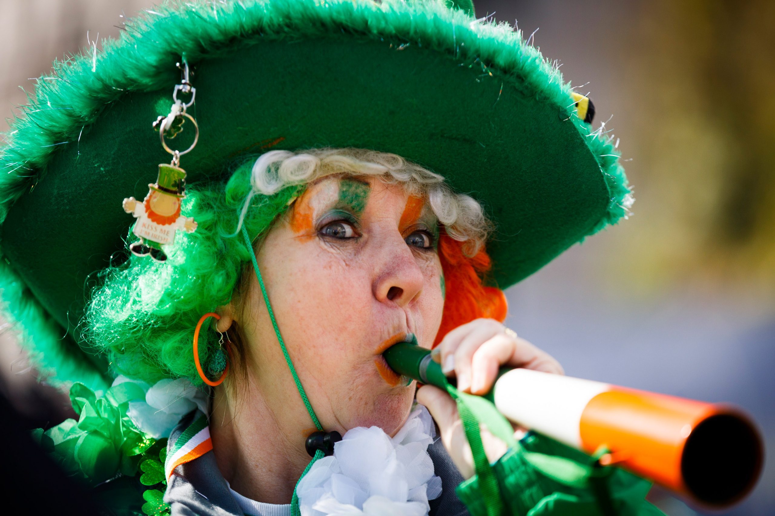Woman blowing horn, dressed in festive green attire, with a large hat and matching face paint, amidst outdoor celebrations. Keychain reads "Kiss Me I'm Irish."