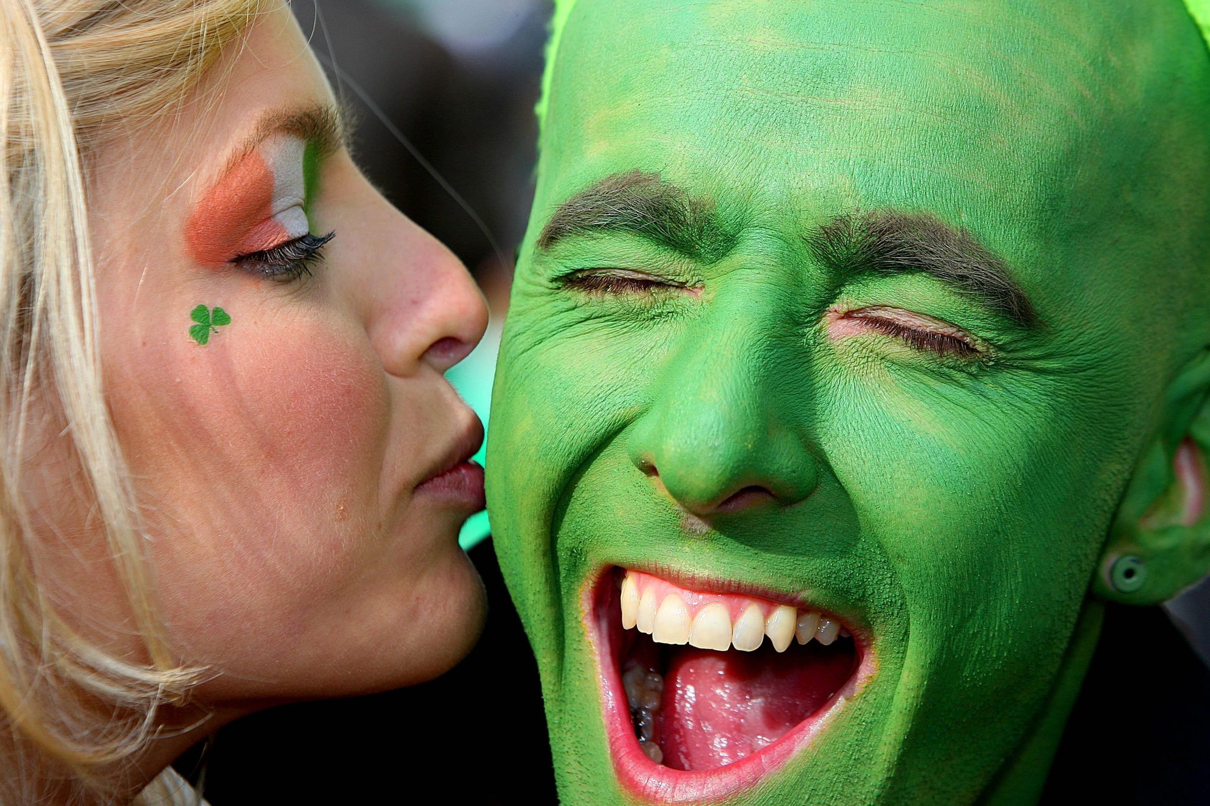 Face painted green laughs joyfully, receiving a cheek kiss from a woman with shamrock face paint, celebrating in a lively, festive atmosphere.