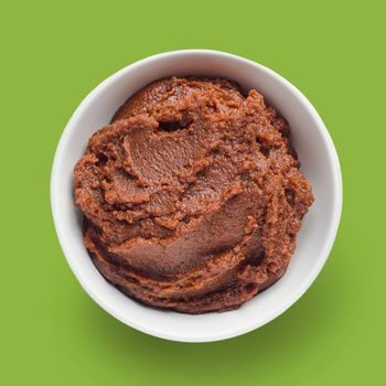 A white bowl holds brown paste, resting on a solid green background.