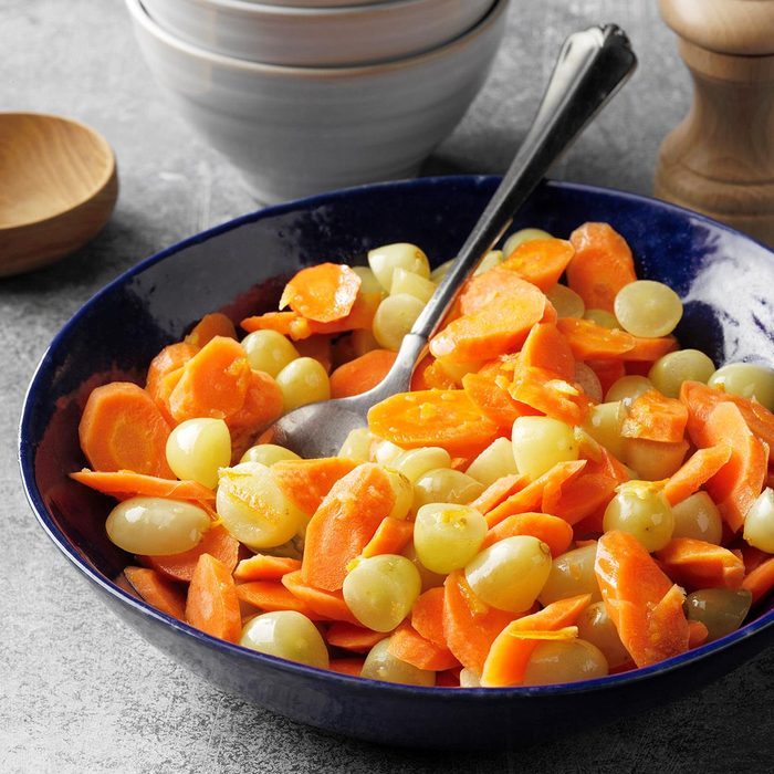 Bowl holds sliced carrots and grapes, mixed together with a spoon, on a gray countertop, surrounded by stacked bowls and a pepper grinder.