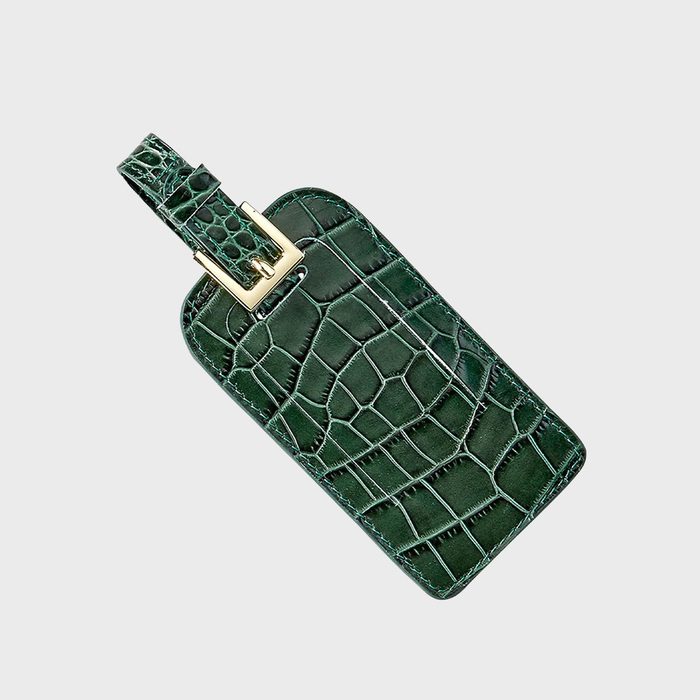 Green crocodile-patterned luggage tag lies diagonally, featuring a gold buckle, on a plain light gray background.