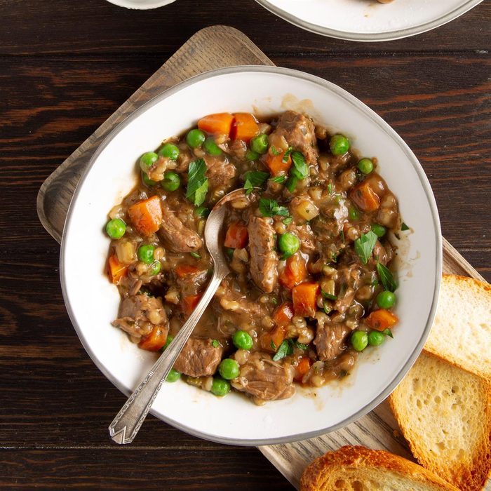Stew sits in a white bowl with a spoon, featuring beef, peas, and carrots; accompanied by slices of bread on a wooden table.