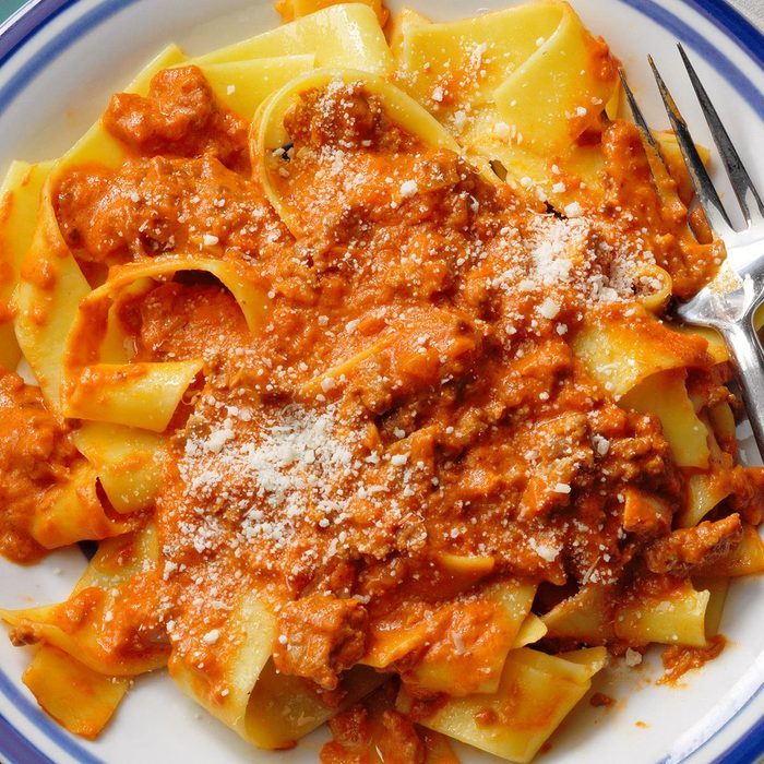 Pasta topped with rich meat sauce and sprinkled with cheese, resting on a white plate with a blue rim, alongside a metal fork.