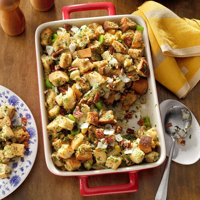 Stuffing sits in a red baking dish, resting on a wooden table, garnished with herbs and pecans, alongside a plate, spoon, and yellow napkin.