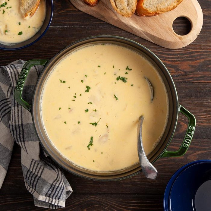 Soup sits steaming in a green pot, garnished with herbs, on a dark wooden table beside sliced bread and a plaid cloth.