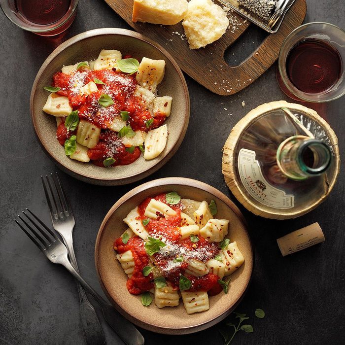 Bowls of gnocchi topped with tomato sauce and basil sit alongside a bottle of wine, glasses, and cheese on a dark table.