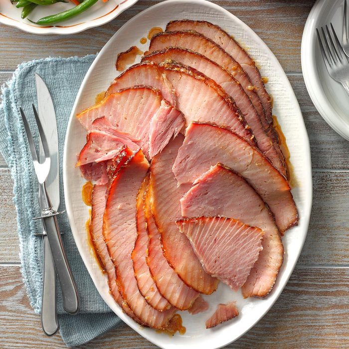 Slices of glazed ham are arranged on a white platter, accompanied by a knife and fork on a blue napkin, with green beans nearby.