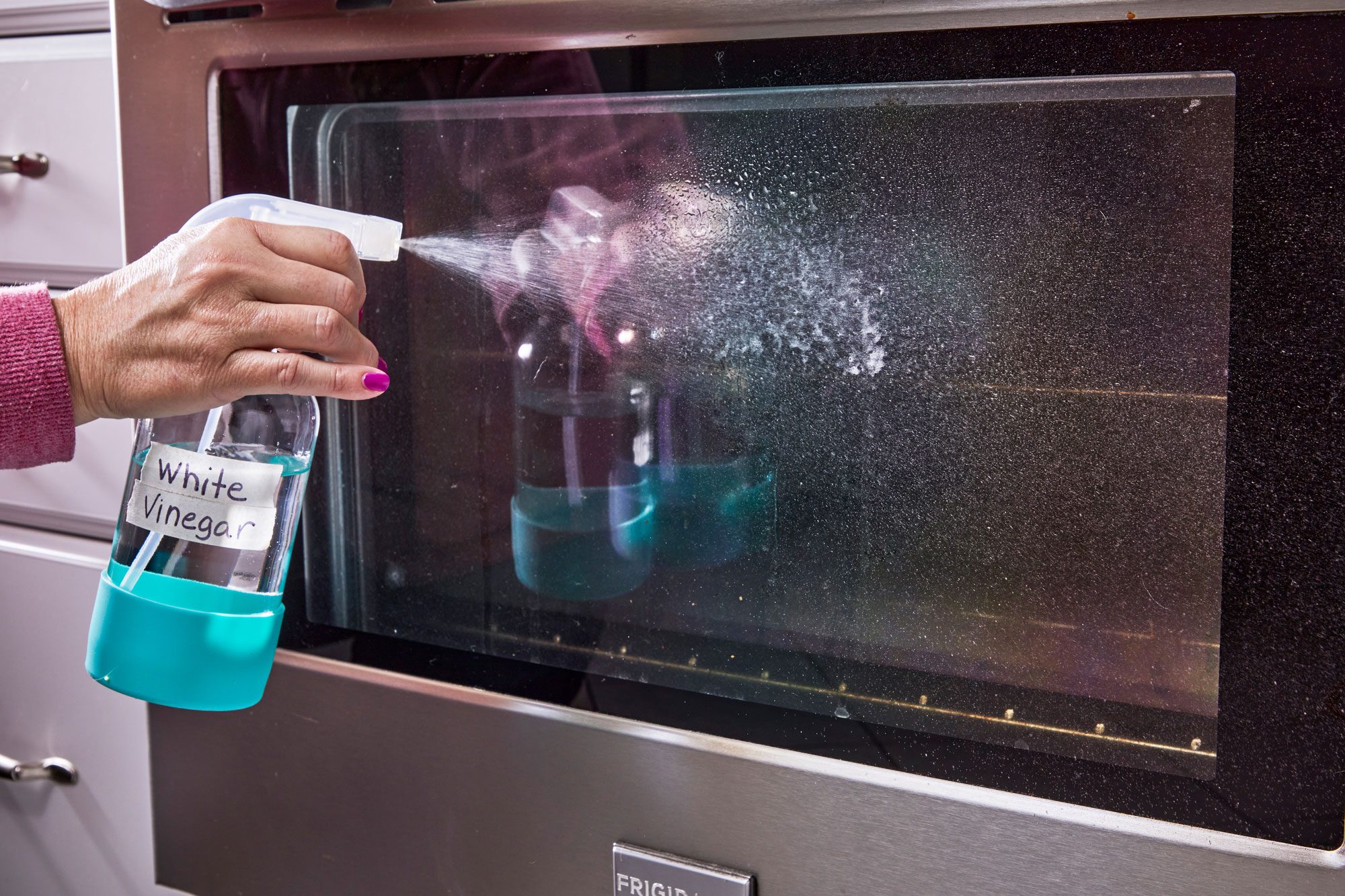 hand spraying white vinegar onto exterior of glass oven door