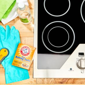 Cleaning supplies and baking soda are arranged beside a ceramic stovetop. A scraper, gloves, cloths, and vinegar complete the setup on a wooden surface.