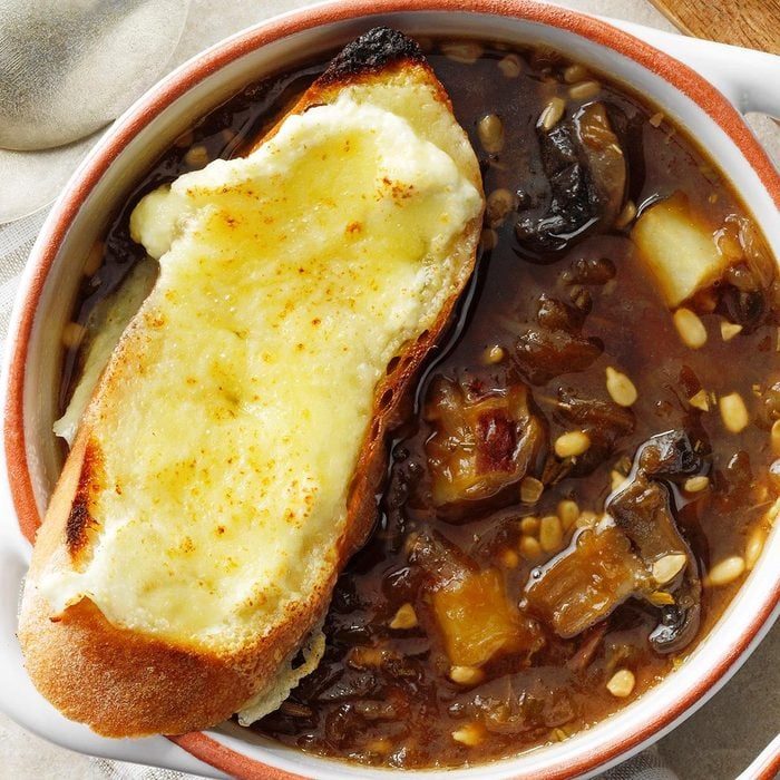 A bowl containing onion soup and a slice of crusty bread topped with melted cheese; the table features a white cloth.