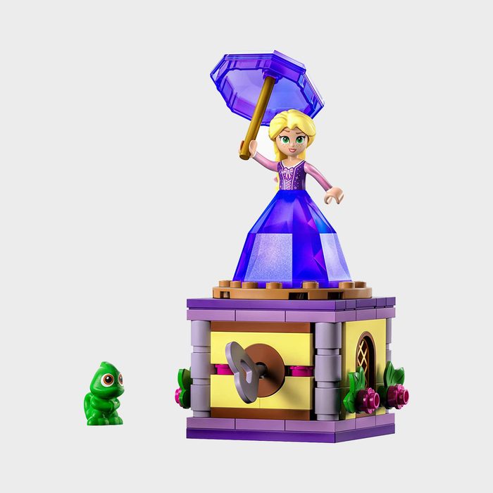 A LEGO figure twirls with umbrella on a decorated box platform, observed by a small green chameleon, on a plain background.