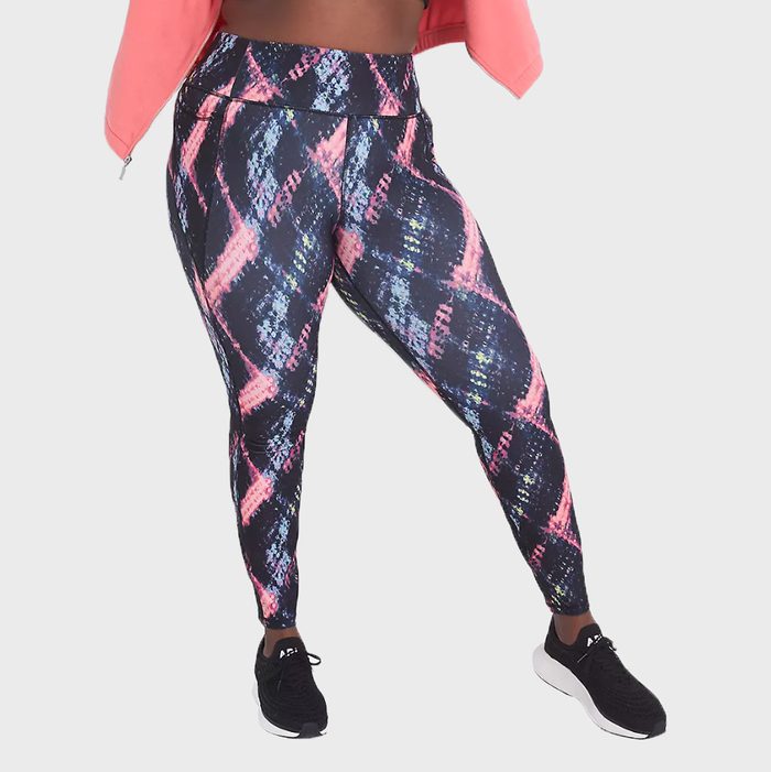 Leggings display vibrant geometric pattern. Person stands confidently with legs apart, wearing black sneakers. Background is plain and neutral.