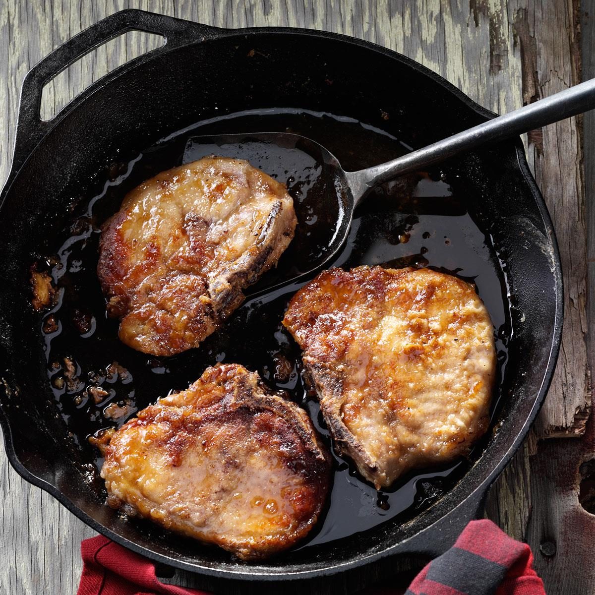 Pork chops sizzle in a cast-iron skillet, sitting on a weathered wooden table with a red and black checked cloth nearby.