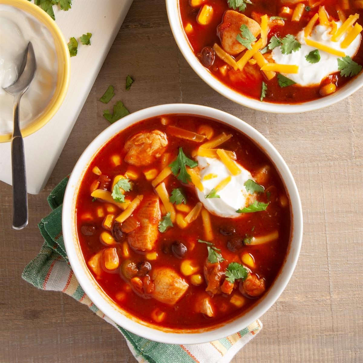 Two bowls of chili topped with cheese and sour cream sit on a wooden table, accompanied by a spoon and napkin.