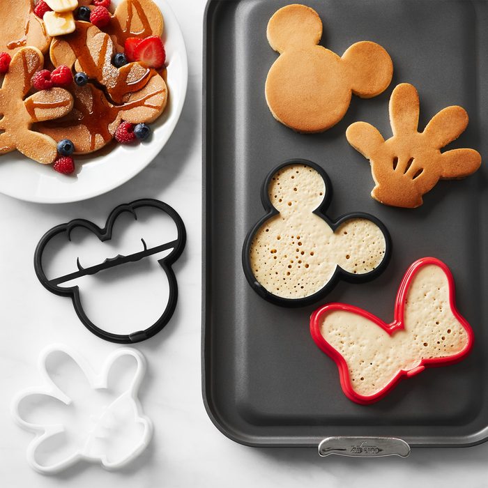 Mickey and Minnie pancake molds frame batter on a griddle; cooked pancakes with berries and butter rest on a nearby white plate.