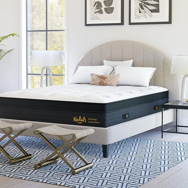 Mattress sits on an upholstered bed frame, surrounded by two stools, a lamp, and abstract art in a bright, modern bedroom. Text reads 