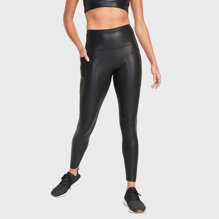 Person wears shiny black leggings and a cropped top, standing confidently with one hand in pocket, against a plain gray background.