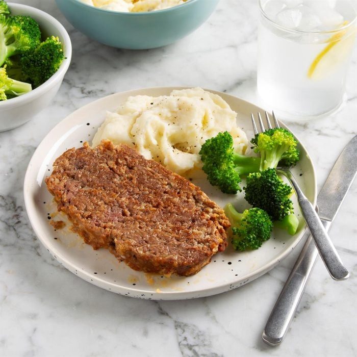A meatloaf slice sits on a plate with mashed potatoes and broccoli, accompanied by fork and knife, on a marble surface.