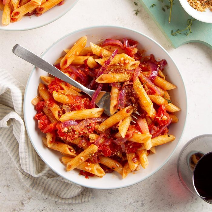 Bowl of penne pasta mixed with tomato sauce and onions, sprinkled with cheese, on a white countertop with a striped cloth and a glass of wine.