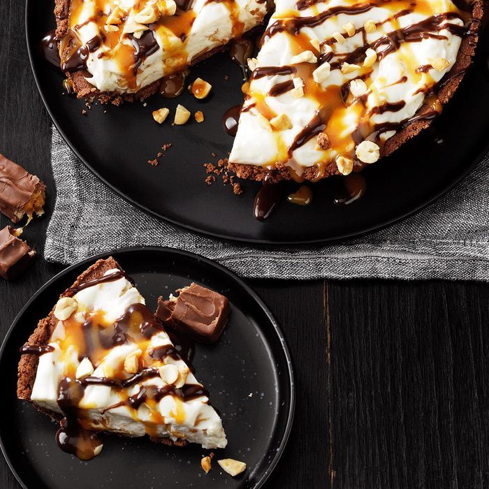 Pie slice drizzled with caramel and chocolate, garnished with nuts, rests on a black plate alongside a chocolate piece on a dark wood table.