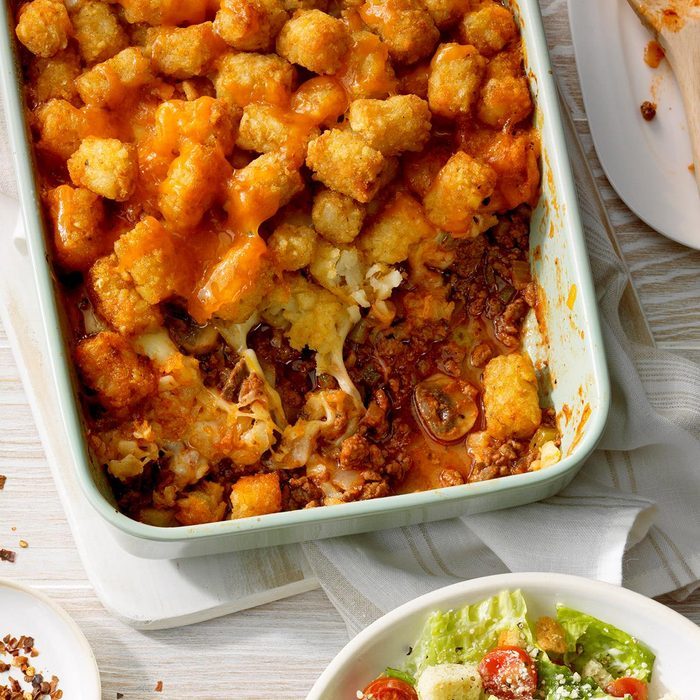 Casserole displays crispy tater tots atop melted cheese and ground meat in a baking dish, accompanied by a fresh salad on a light wooden table.