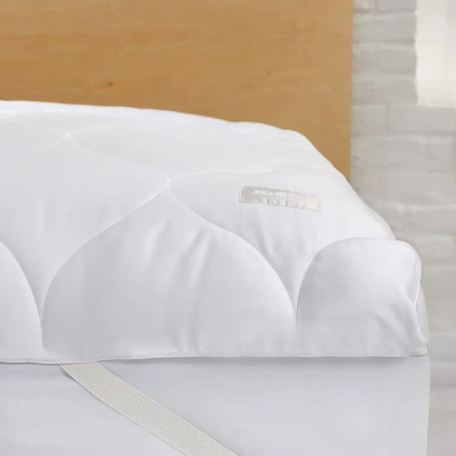 Mattress topper rests on bed; white, quilted, with strap securing it; brick wall and wooden headboard in the background.