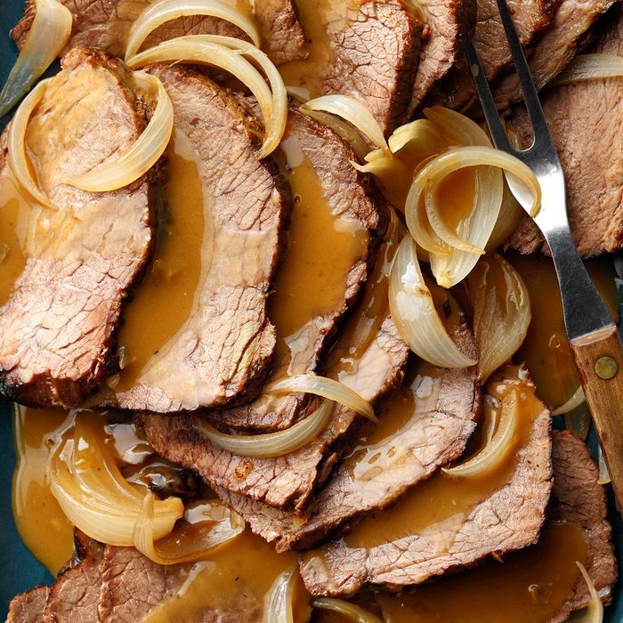 Sliced roast beef covered in gravy, topped with caramelized onions, rests on a blue plate, accompanied by a carving fork.