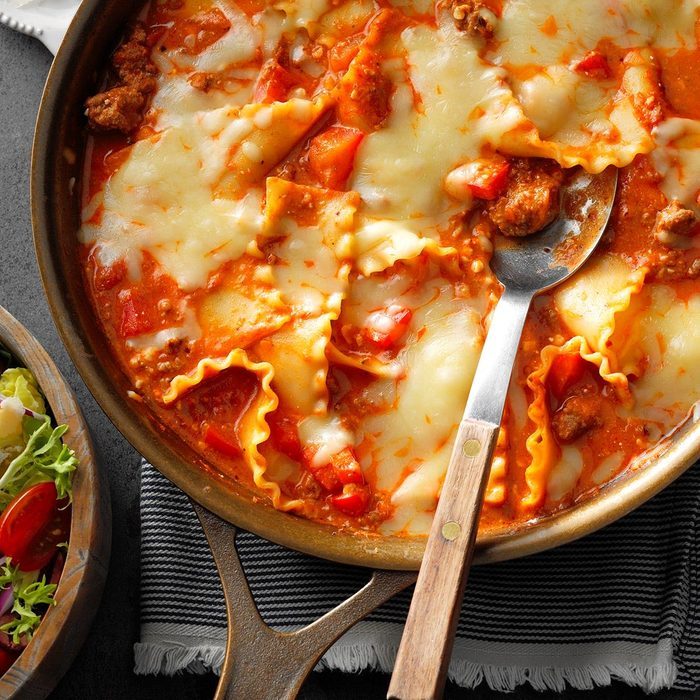 Skillet lasagna bubbles in a pan, covered with melted cheese, near a salad bowl, placed on a striped cloth on the table.