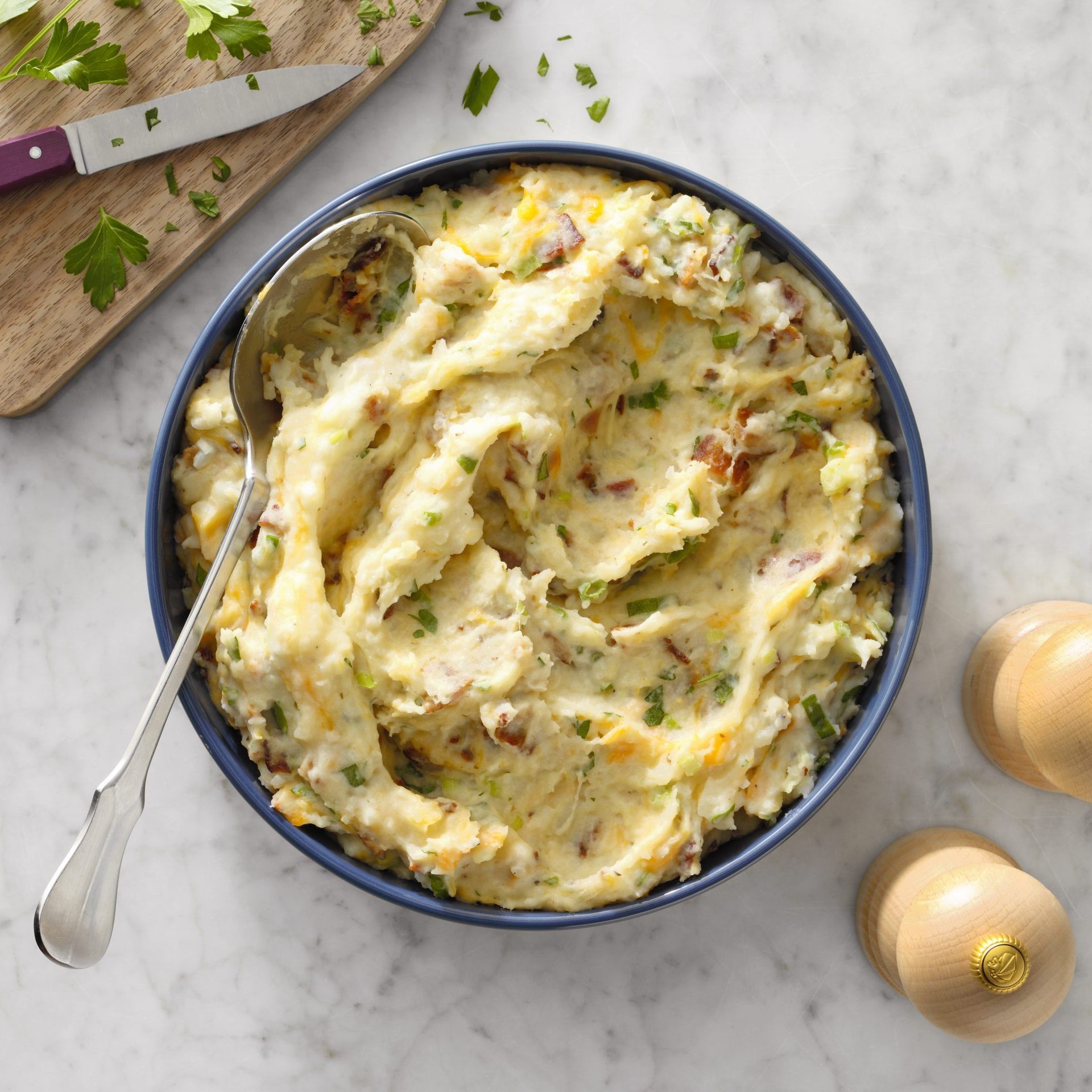 Mashed potatoes in a blue bowl with a spoon, garnished with herbs. Chopped parsley and a knife on a wooden board are nearby.