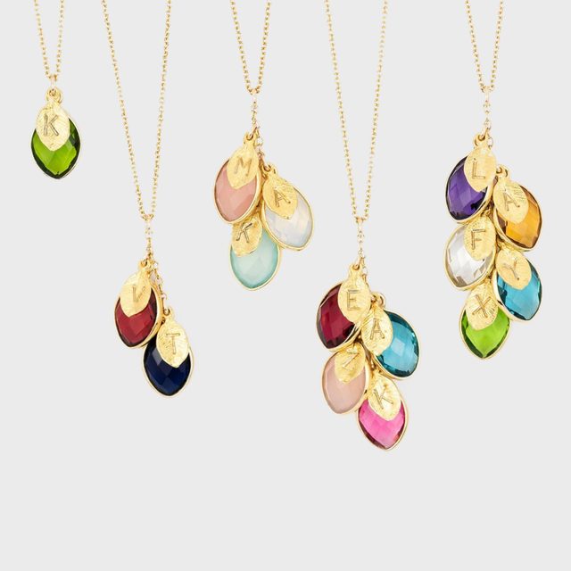 Necklaces display colorful teardrop stones and gold charms engraved with letters. Positioned against a plain background, chains hang vertically displaying pendants in groups.