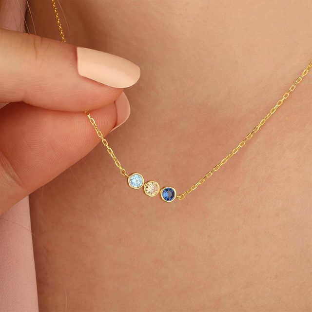 Hand holds a golden necklace with three gemstones: light blue, clear, and dark blue. The necklace rests against skin.