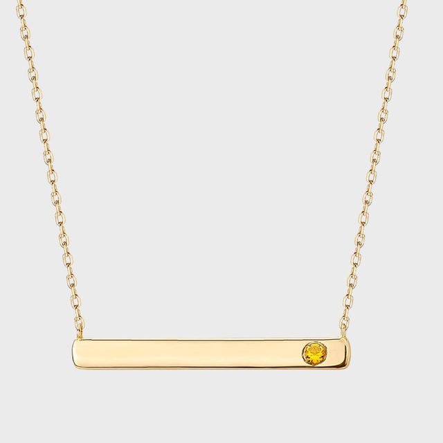 Gold bar necklace with a small yellow gemstone on one side, hanging from a delicate chain against a plain background.