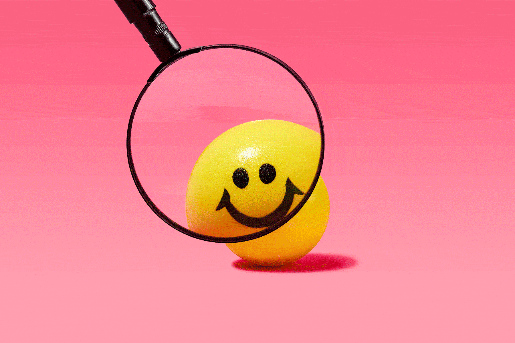 Magnifying glass magnifying different angles of a yellow smiley face ball to investigate what is happiness; pink background
