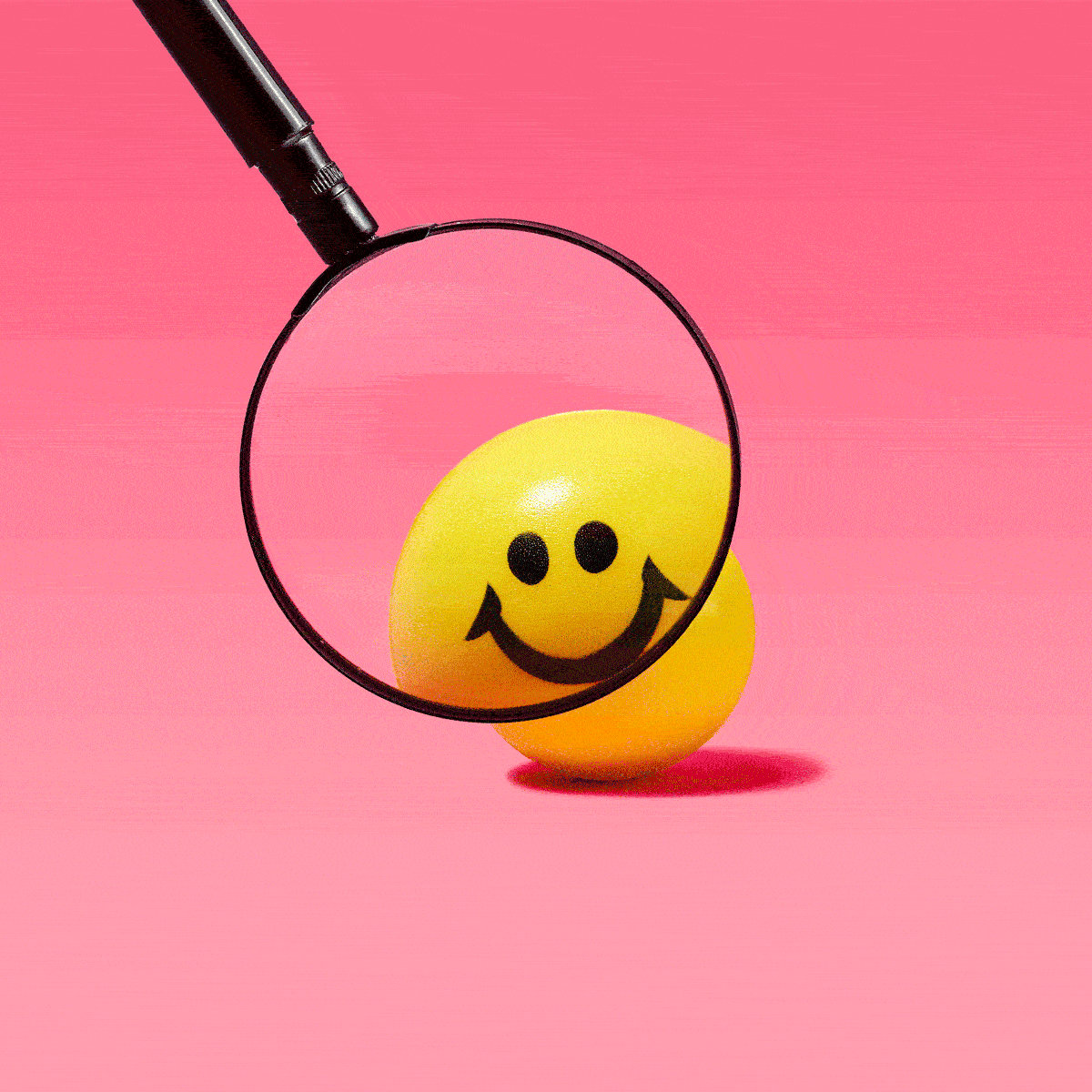 Magnifying glass enlarges a yellow smiley face ball against a pink background.