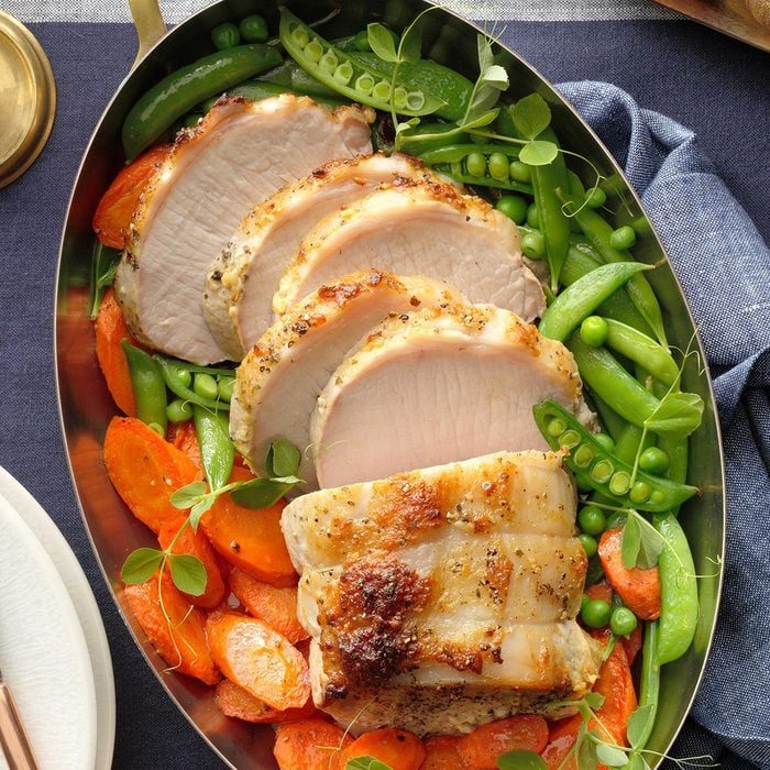 Sliced roasted pork sits atop a bed of vibrant green peas and snap peas, with seasoned orange carrots, in an oval metal dish.