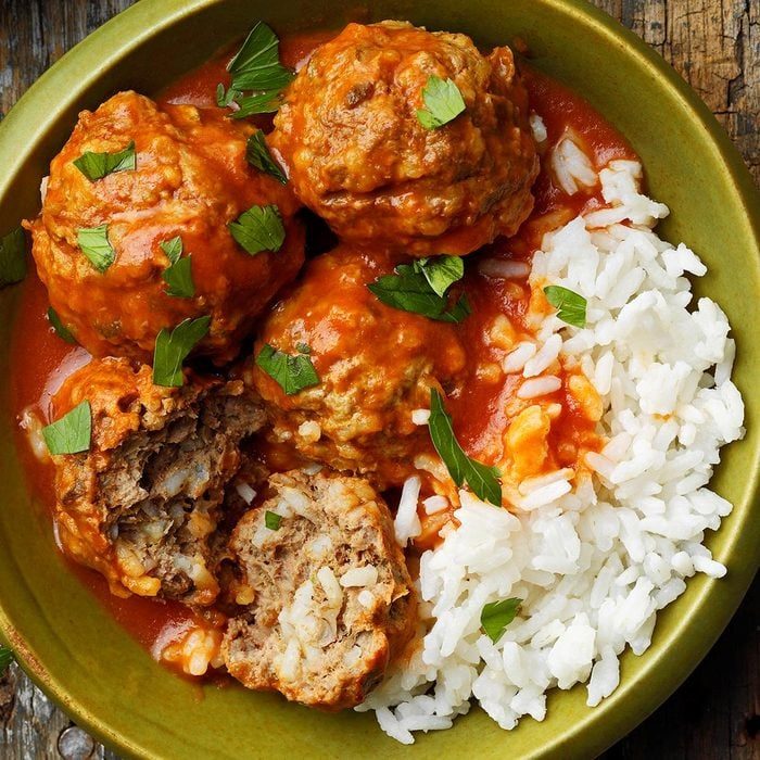 Meatballs sit in tomato sauce, garnished with herbs, next to white rice in a green bowl on a wooden surface.