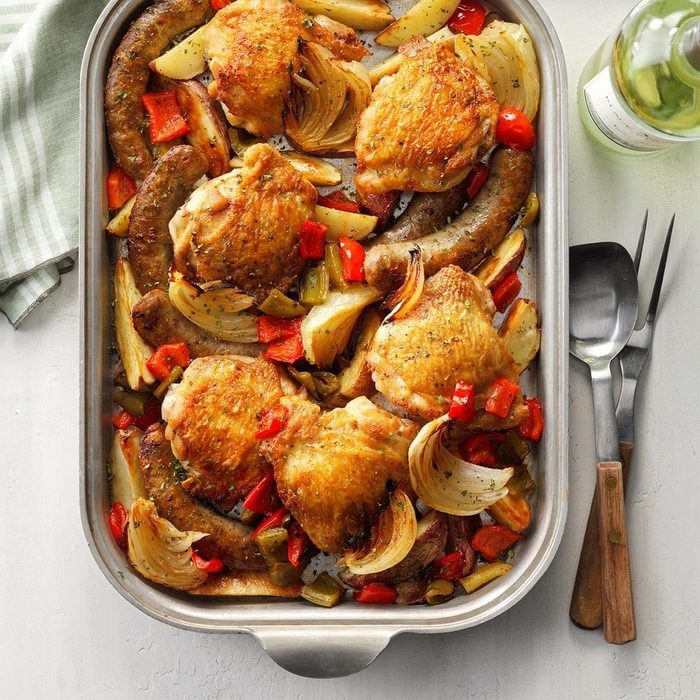 Baked chicken and sausages rest with roasted onions and peppers in a large metal baking dish on a white table, next to a green-striped cloth.