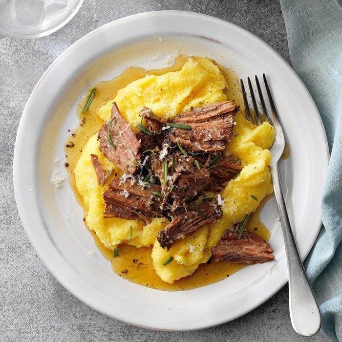 Shredded beef rests on creamy polenta, garnished with herbs and grated cheese, served on a white plate with a fork nearby on a gray table.