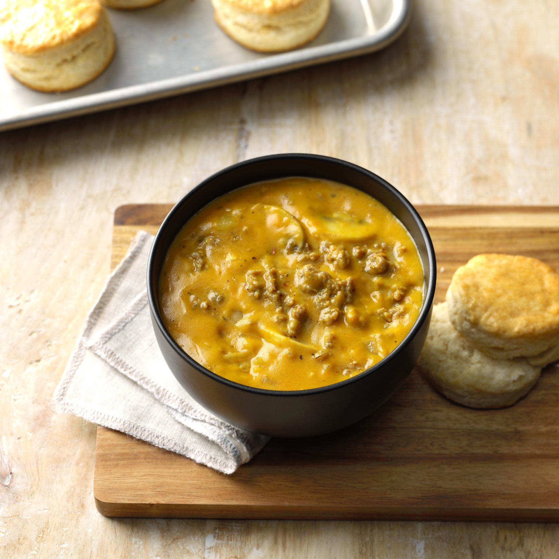 Bowl of thick soup sits on a wooden board, accompanied by two biscuits, with additional biscuits on a baking tray in the background.