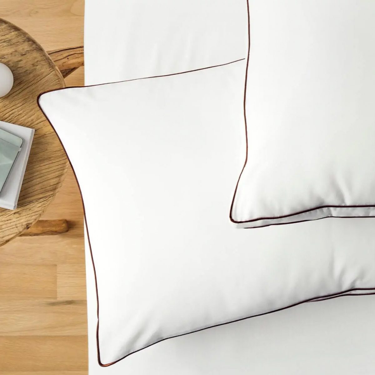 White pillows rest on a bed, accented with brown edges. Nearby, a round wooden table holds a small white plate and phone.
