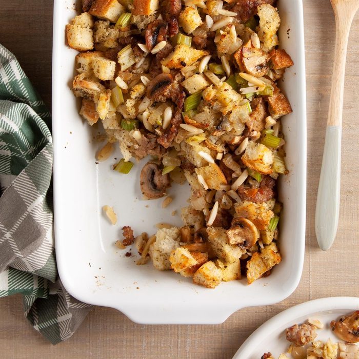 Baking dish holding savory stuffing with bread cubes, mushrooms, and celery; partially served onto an adjacent white plate on a wooden table.