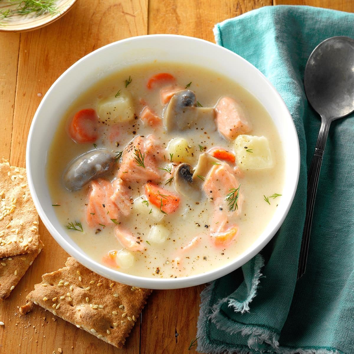 Bowl contains creamy soup with salmon, mushrooms, carrots, and potatoes; linen napkin and spoon on wooden table, sesame crackers beside.