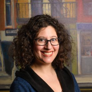 Person smiling, wearing glasses and a dark jacket, stands indoors with a colorful blurred painting in the background.