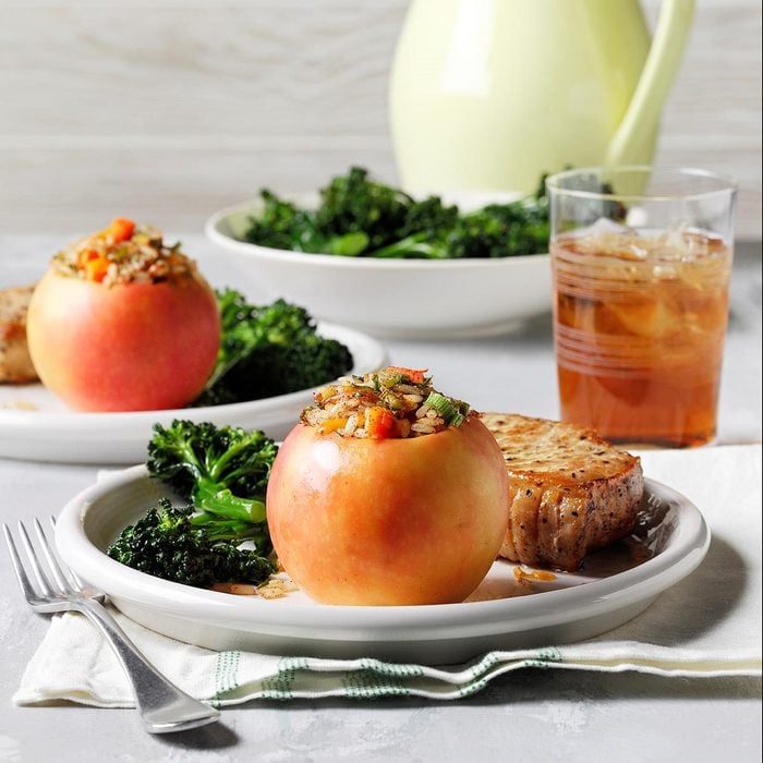 Stuffed apple sits on a plate with broccoli and toast; iced drink and second plate in background on a light-colored table.