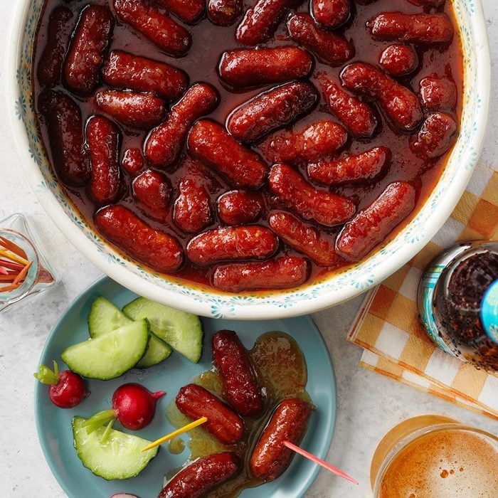 Sausages in sauce fill a floral bowl; nearby, a plate holds cucumbers, radishes, and toothpicked sausages on a checkered napkin.