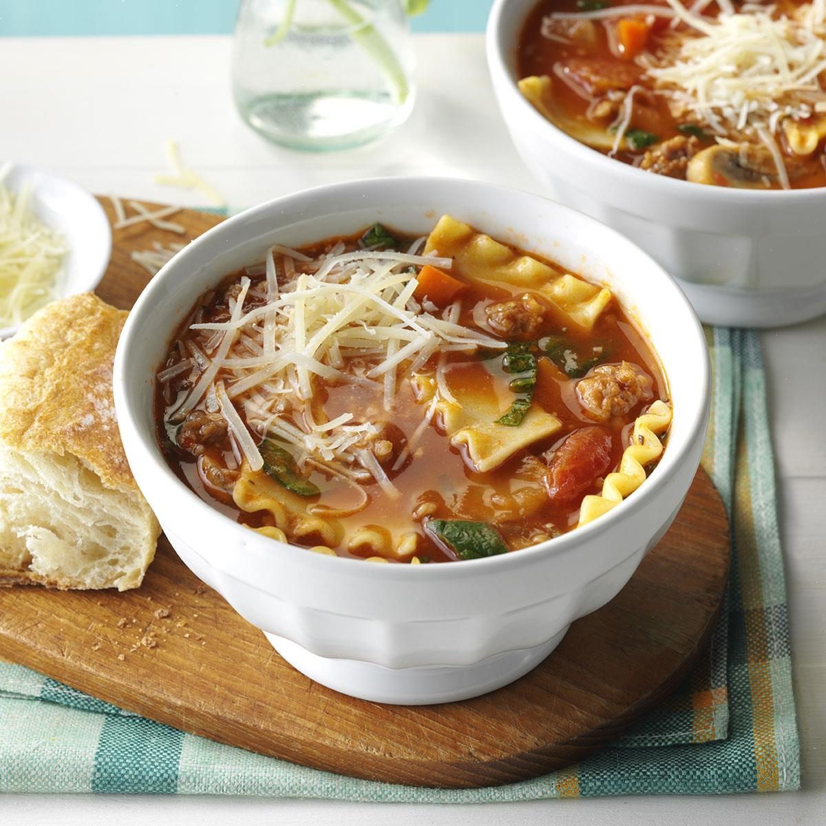 Soup filled with pasta, vegetables, and cheese sits steaming in a white bowl. A crusty bread piece rests on a wooden board nearby.