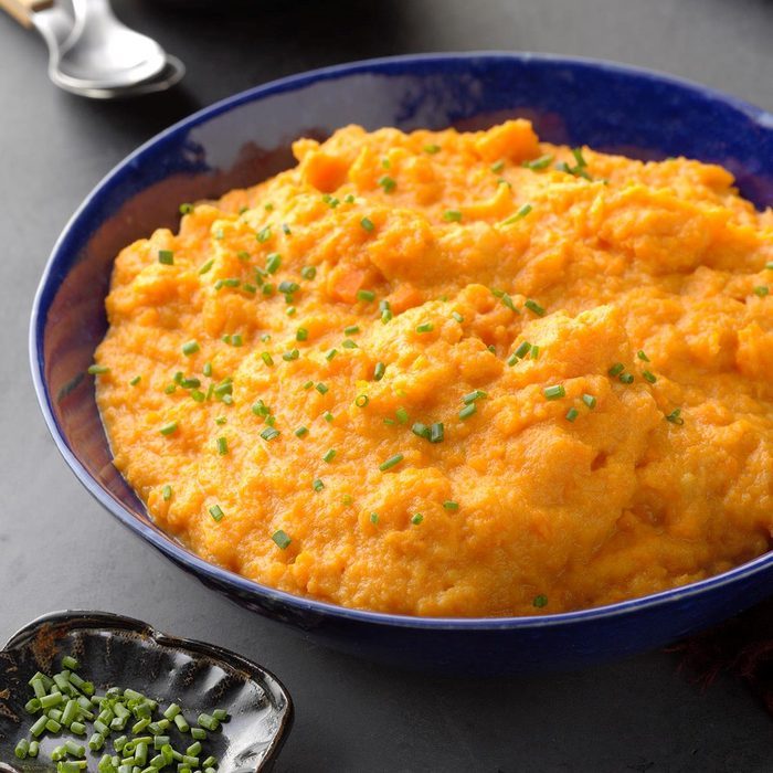Mashed sweet potatoes, garnished with chopped chives, sit in a blue bowl on a dark surface, with extra chives in a small dish nearby.